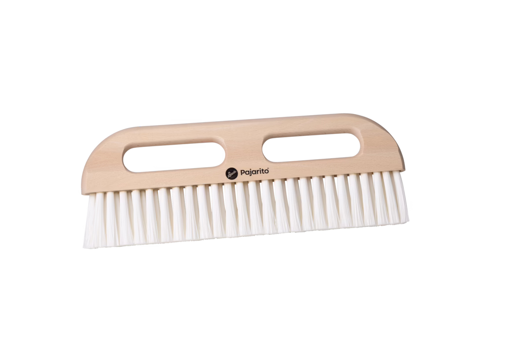 Pajarito_Dummy_Wallpaper squeegee nylon P-Line_179112_2024-02 Productfoto van een houten handborstel met twee langwerpige ovale gripopeningen en een glad, licht beukenhouten handvat. De borstel heeft dichte, rechte witte kunststofharen die in een doorlopende rij langs de onderrand zijn geplaatst. Het kleine zwarte 'Pajarito'-logo is in het midden van het handvat afgedrukt. Gefotografeerd op een witte achtergrond met zacht, gelijkmatig licht, in horizontale positie; geschikt om in huis te vegen en stof te verwijderen.