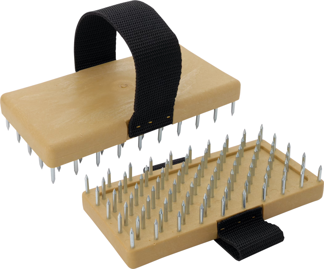 Two rectangular beige plastic blocks fitted with multiple short, pointed metal spikes arranged in neat rows; each block has a wide black woven strap looped over the top and fastened with visible stitching, forming a handle for gripping. The spikes are evenly spaced and slightly tapered to sharp points, made of shiny galvanized metal, and set into the molded plastic base which shows subtle texture and rounded corners. The image shows one block positioned above the other at an angle so the underside with the spikes is visible on both, highlighting the regular pattern of the pins and the functional, hand-held design. The overall impression is of a compact, durable handheld tool or accessory with a utilitarian aesthetic, suitable for tasks requiring repeated contact with a surface.