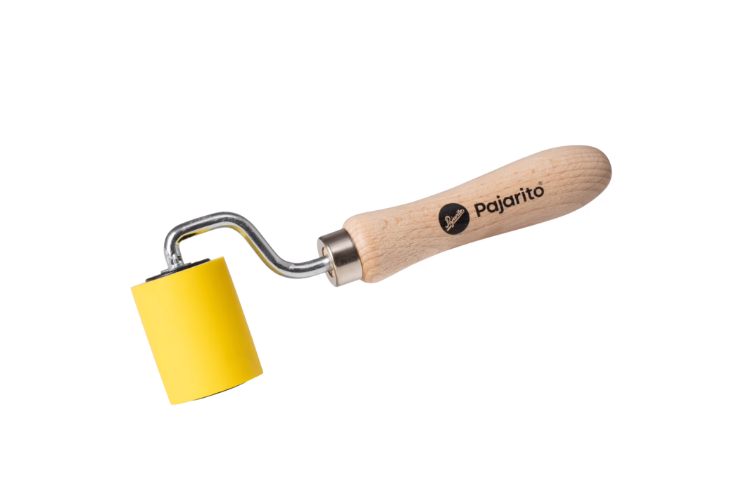 High-resolution product photograph of a small hand roller tool photographed on a clean white background. The tool features a smooth, light beech-wood handle printed with the brand name "Pajarito" and a round logo in black. A chrome-plated metal shaft with a distinctive bent offset connects the handle to a cylindrical yellow rubber or foam roller. The roller is bright yellow, approximately small-to-medium in diameter, with flat ends and a uniform surface. The composition places the tool diagonally across the frame from lower left toward upper right, showing clear detail: visible wood grain and texture on the handle, subtle metallic reflections on the shaft, and soft shadowing beneath the roller and handle. The lighting is even and neutral, emphasizing the product’s colors and materials without harsh contrasts. This image is suitable for e-commerce, product catalogs, or instructional content and conveys the tool’s size, materials, branding, and intended use for smoothing, pressing or applying even pressure on surfaces such as wallpaper, vinyl, adhesives or printmaking applications.
