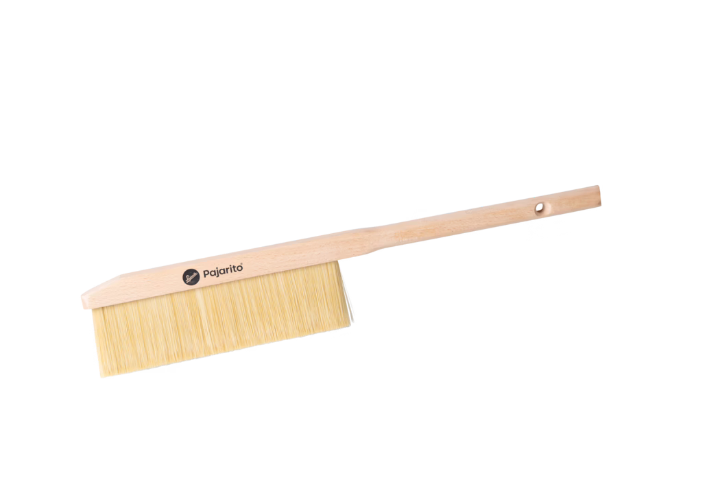 Close-up of a long-handled wooden hand brush photographed on a white background. The brush features a light beech wood handle and a slim rectangular head fitted with dense, pale natural bristles. A small round logo with the word "Pajarito" is printed on the wooden head, and a drilled hanging hole is visible near the end of the handle. The simple, utilitarian design suggests the brush is intended for indoor sweeping or dusting of surfaces and workbenches.
