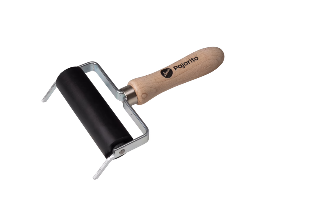 Pajarito_Dummy_Pressure roller support_036300_2021-02 Een kleine handroller (brayer) op een witte achtergrond. Hij heeft een zwarte rubberen roller in een gebogen metalen frame en een ergonomisch licht houten handvat met de opdruk 'Pajarito'. Het metalen frame zit vast met zichtbare klinknagels; het houten handvat toont natuurlijke houtnerf en een afgerond uiteinde. Het gereedschap wordt gebruikt om inkt of verf gelijkmatig te verdelen op drukplaten, linoleum of papier en ziet er schoon en ongebruikt uit. De compositie is licht diagonaal met de roller linksonder en het handvat naar rechtsboven.