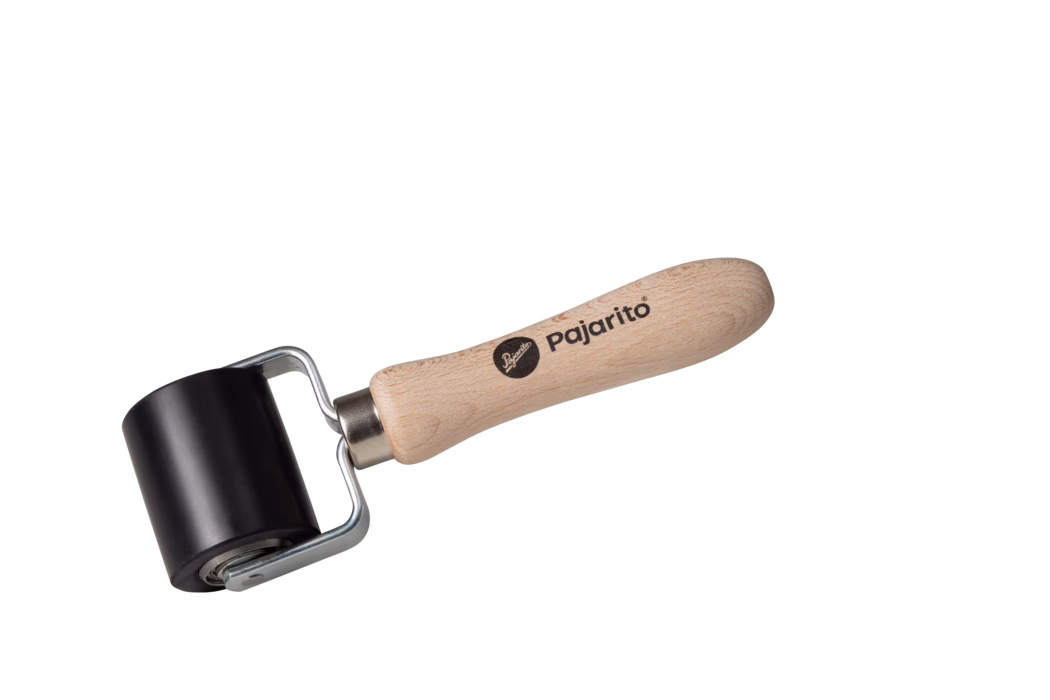 A small hand roller tool photographed on a white background, consisting of a smooth black rubber cylindrical roller mounted on a metal frame and attached to a light beech wood handle stamped with the brand 'pajarito'. The handle is ergonomically shaped, sanded, and shows visible wood grain; the metal bracket connects to a bearing allowing the roller to rotate freely. The tool is isolated, well-lit, and positioned diagonally with the handle on the right and the roller on the left. Suitable for tasks such as applying pressure when laminating, smoothing wallpaper or decals, and printmaking.