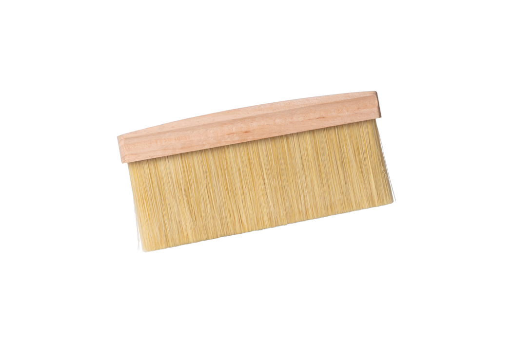 Een brede platte borstel met een licht houten handvat en dicht opeengepakte beige synthetische haren, afgebeeld tegen een witte achtergrond in een lichte diagonale hoek. De borstel wordt doorgaans gebruikt bij het behangen, stofvegen of het aanbrengen van lak; de lange, dicht geplaatste haren zijn bevestigd aan een eenvoudige onbehandelde houten rug.